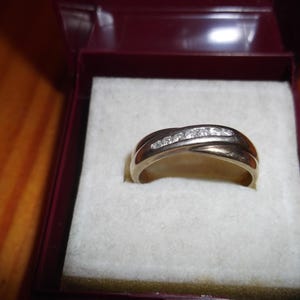 Può includere: Un anello dorato con una fila di piccole pietre trasparenti sulla parte superiore. L'anello è presentato all'interno di un portagioie bordeaux con interno bianco. L'anello ha un design curvo.