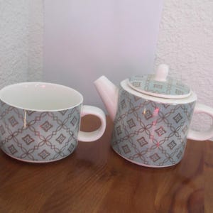 May include: A ceramic tea set featuring a cup and a teapot. Both pieces are light blue with a repeating geometric pattern in a darker shade of blue and brown. The teapot has a lid and a spout.