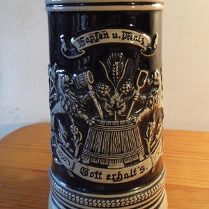 Peut inclure: Une chope à bière en céramique bleu foncé avec un motif en relief détaillé. La chope présente du texte et des images de houblon, d'orge et d'un tonneau. Les bords supérieur et inférieur sont blancs avec un motif décoratif.