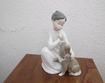 May include: A ceramic figurine of a child in a white dress, seated and interacting with a small dog. The child has gray hair and is touching the dog, which is brown and white. The figurine is on a wooden surface.