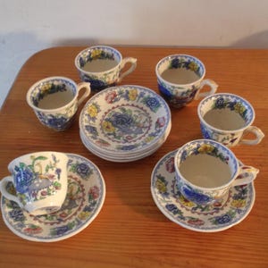 Puede incluir: Un juego de seis tazas y platillos de té vintage. Las tazas y los platillos son blancos con un diseño floral en azul, amarillo y rojo. Las tazas tienen asas pequeñas y los platillos son redondos.