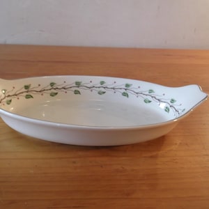 May include: An oval serving dish in white, decorated with a vine and leaf pattern. The rim is trimmed with a gold-coloured band. The dish has handles at each end.