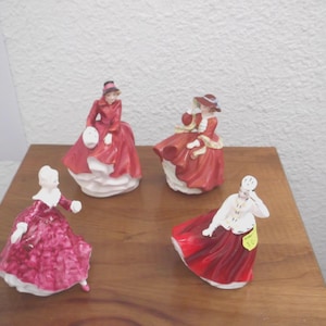 May include: Four porcelain figurines of women in vintage dresses. The figurines are in shades of red, white, and pink, with detailed clothing and accessories. They are posed in various stances, suggesting movement and elegance.