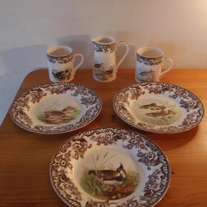 Puede incluir: Un juego de vajilla de cerámica vintage con diseños de patos, peces y flores. El juego incluye tres platos, dos tazas y una jarrita. Los platos y tazas tienen un borde floral marrón. La jarrita tiene un asa.
