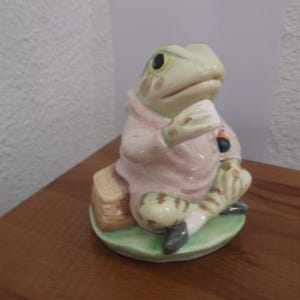 Op de afbeelding: Een keramieken kikkerfiguur in zittende positie, gekleed in een roze hoodie. De kikker heeft een lichtgroen lichaam met donkere ogen en zit op een groene basis. Het beeldje staat op een houten oppervlak.