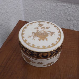 May include: A round white porcelain trinket box with a lid featuring a gold floral basket design. The side of the box has a dark blue band with gold scrollwork and gold floral accents. The base is white with gold floral patterns around the perimeter.
