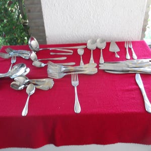 May include: A large collection of silver-toned stainless steel flatware arranged on a red surface. The set includes various forks, spoons, knives, a ladle, and a cake server, all featuring a simple, smooth handle design with rounded ends.