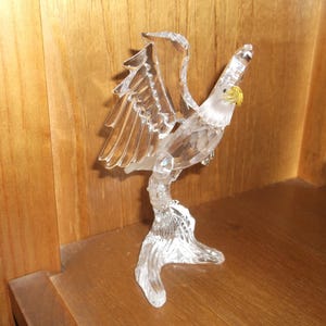 May include: A clear crystal eagle figurine with outstretched wings and a yellow beak. The eagle is perched on a clear crystal base, and the wings are detailed with individual feathers. The figurine is displayed on a wooden surface.