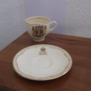 Könnte beinhalten: Ein Vintage-Tee-Set, bestehend aus einer Tasse und einer Untertasse. Die cremefarbene Tasse hat einen Henkel und ein dekoratives Emblem. Die Untertasse ist cremefarben mit einem Goldrand und einem Kronen-Emblem mit den Buchstaben "ER".