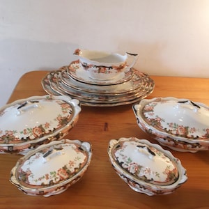 May include: A collection of vintage porcelain serving dishes and plates. The set includes several oval covered dishes, a gravy boat, and multiple plates. The dishes feature a floral pattern with gold trim, set on a wooden table.