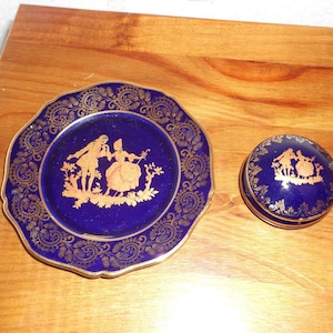 Pode incluir: Um prato decorativo azul cobalto e uma pequena caixa redonda para bugigangas. Ambos os itens apresentam um design dourado de um casal em um cenário de jardim. O prato tem uma borda ondulada e uma moldura dourada ornamentada. A caixa tem uma moldura dourada correspondente.