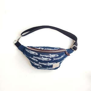 May include: A blue fanny pack featuring a white fish pattern, a brown zipper, and a black adjustable strap. A small rectangular label with the text 'tarrio' is stitched onto the lower right corner of the bag, which includes a metal buckle and hardware.