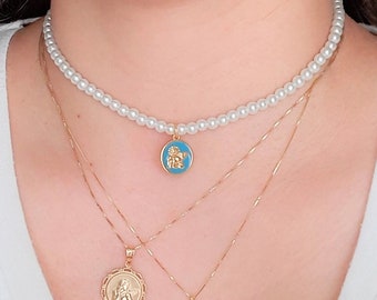 Op de afbeelding: Een gelaagde kettingset met een parelketting met een blauwe en gouden hanger, een gouden ketting met een ronde hanger en een gouden ketting met een kruishanger.