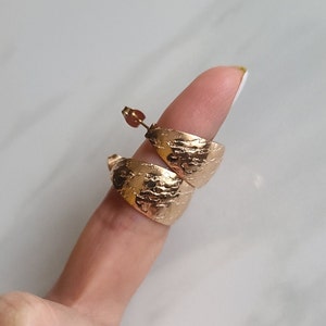 May include: Gold-colored, textured hoop earrings. The earrings have a wide, curved design with a hammered or distressed surface. The earrings are shown on a finger.