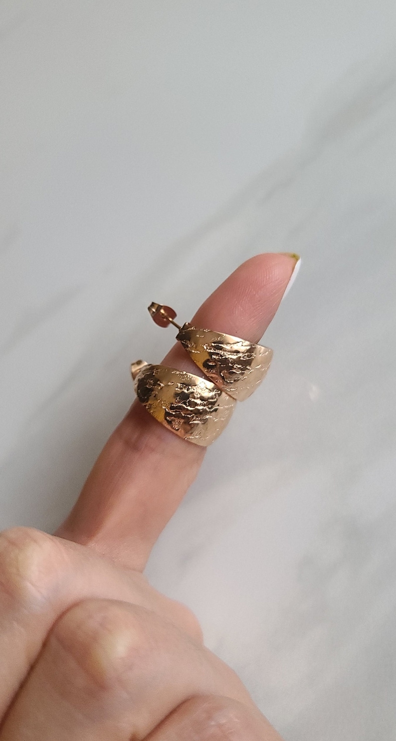 May include: Gold-colored, textured hoop earrings. The earrings have a wide, curved design with a hammered or distressed surface. The earrings are shown on a finger.