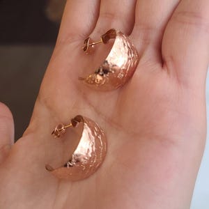 May include: A pair of rose gold crescent moon earrings. The earrings have a hammered texture and are displayed on a hand. The earrings are a warm, metallic color.