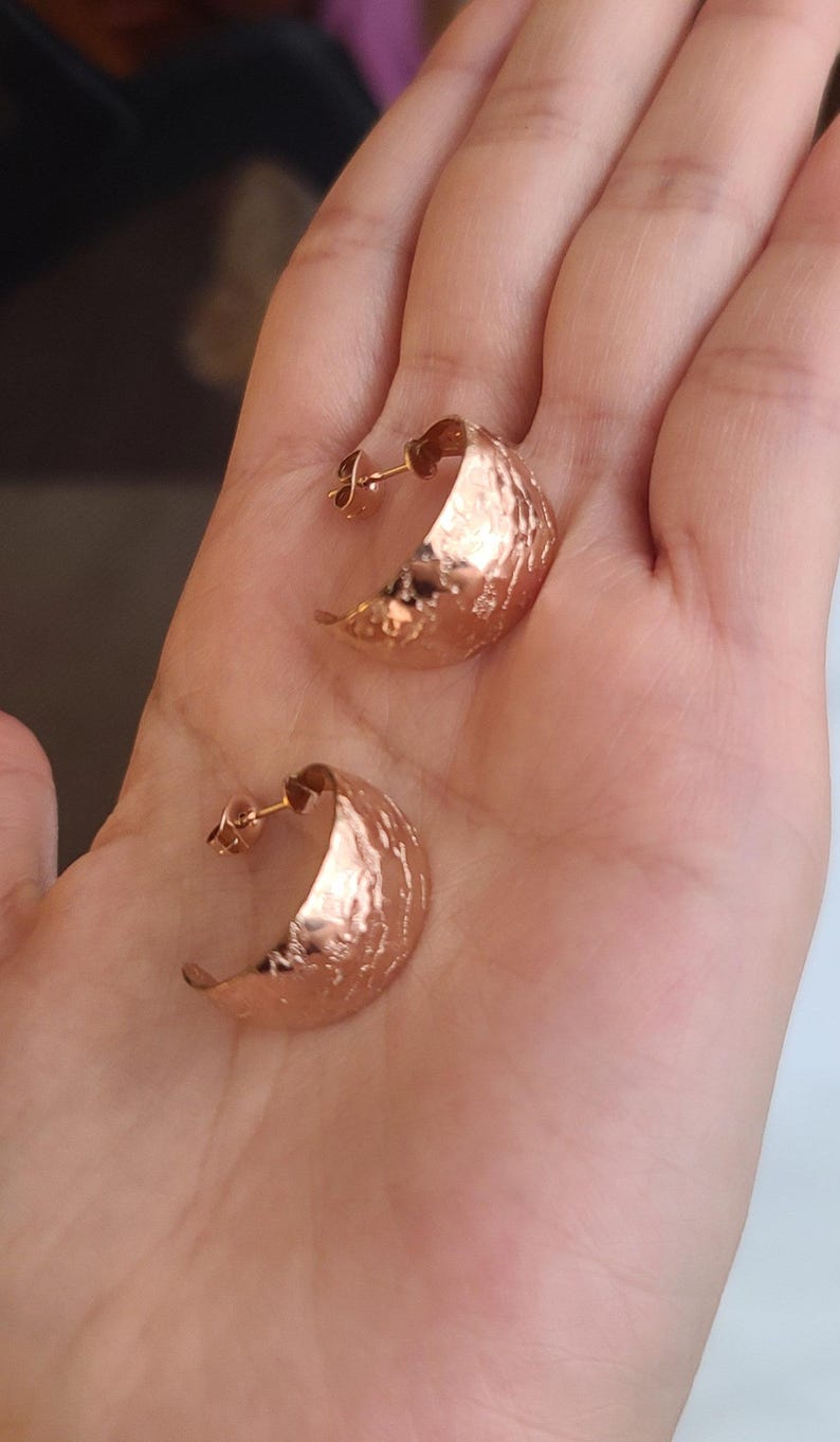 May include: A pair of rose gold crescent moon earrings. The earrings have a hammered texture and are displayed on a hand. The earrings are a warm, metallic color.