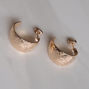 May include: A pair of gold-colored, hammered-texture hoop earrings. The earrings are crescent-shaped with a post backing. The earrings are displayed on a white surface. The earrings are a jewelry item.
