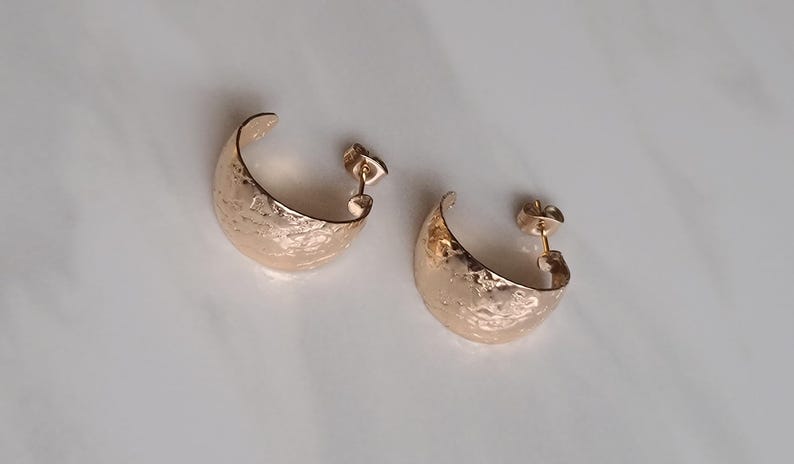 May include: A pair of gold-colored, hammered-texture hoop earrings. The earrings are crescent-shaped with a post backing. The earrings are displayed on a white surface. The earrings are a jewelry item.
