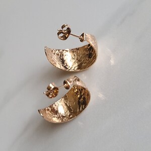 May include: A pair of gold-colored, textured hoop earrings. The earrings have a crescent shape and a post backing. The surface of the earrings has a hammered or textured appearance, reflecting light. The earrings are displayed on a white surface.