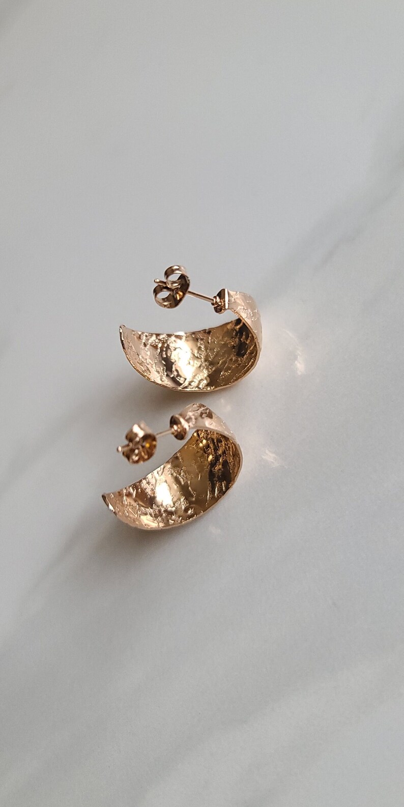 May include: A pair of gold-colored, textured hoop earrings. The earrings have a crescent shape and a post backing. The surface of the earrings has a hammered or textured appearance, reflecting light. The earrings are displayed on a white surface.