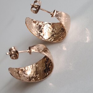 May include: A pair of rose gold hoop earrings with a textured, hammered finish. The earrings have a post and butterfly backing. The earrings are displayed on a white surface.