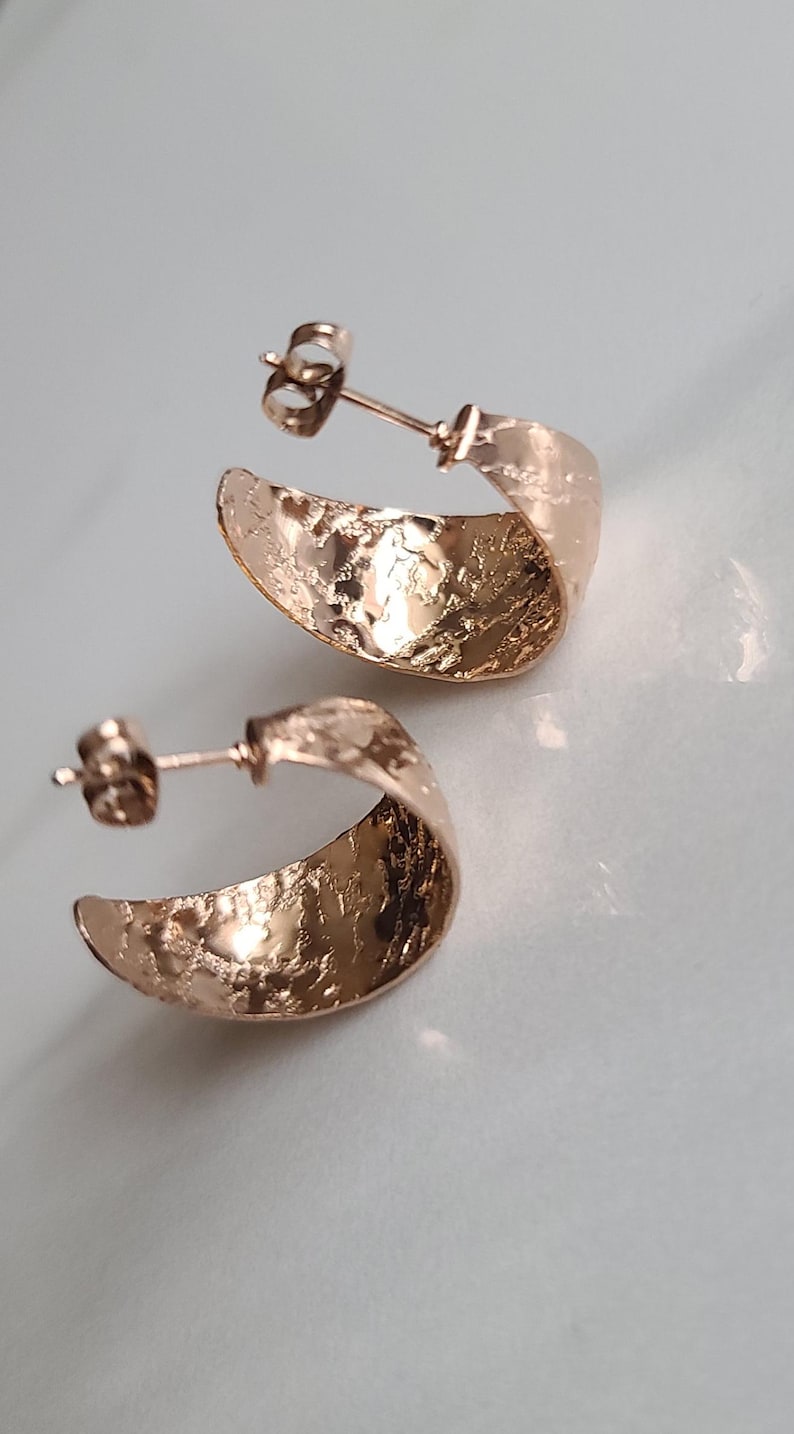 May include: A pair of rose gold hoop earrings with a textured, hammered finish. The earrings have a post and butterfly backing. The earrings are displayed on a white surface.