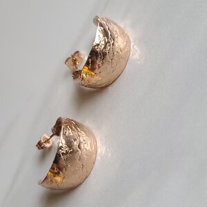 May include: A pair of rose gold half-hoop stud earrings. The earrings have a textured, hammered finish and a post backing. The earrings are displayed on a white surface.