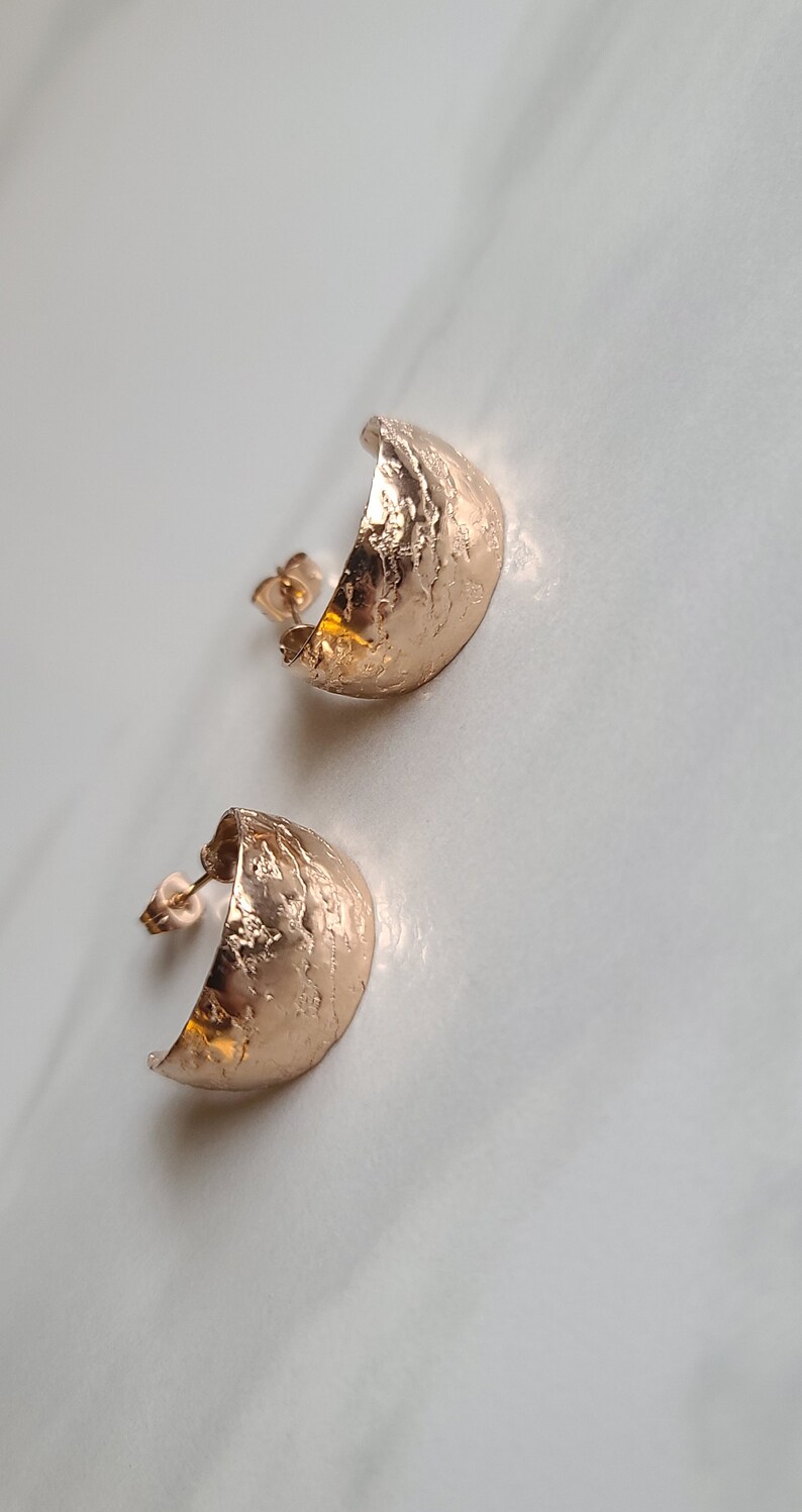 May include: A pair of rose gold half-hoop stud earrings. The earrings have a textured, hammered finish and a post backing. The earrings are displayed on a white surface.