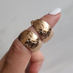 May include: Two gold-colored, textured hoop earrings. The earrings have a hammered or distressed appearance, with a slightly irregular surface. The earrings are displayed on a finger against a neutral background.