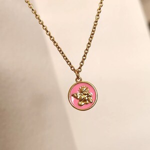 K&ouml;nnte beinhalten: Eine goldene Kettenkette mit einem rosa und goldenen Anh&auml;nger mit einem Cherub-Engel-Design.