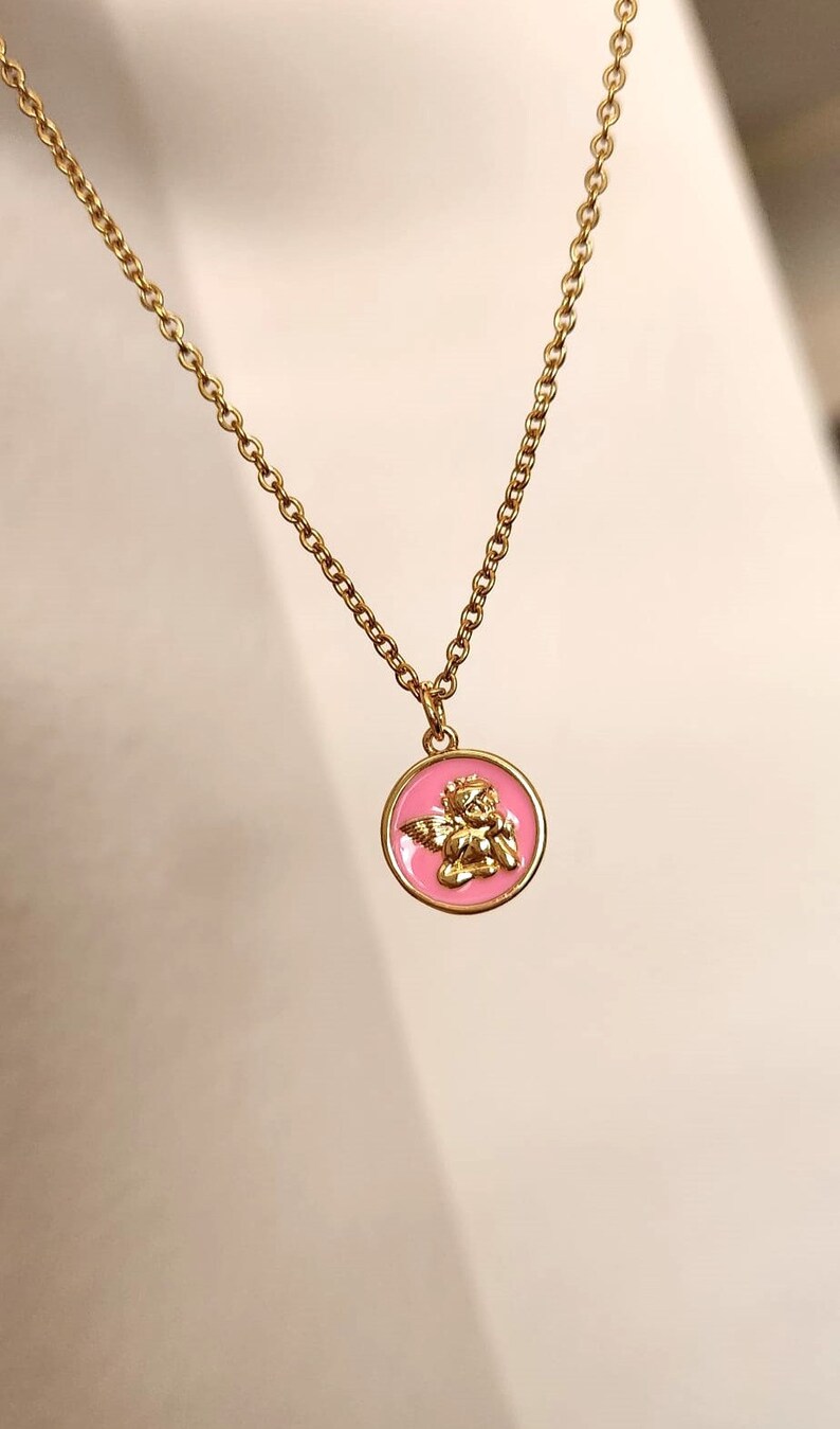 K&ouml;nnte beinhalten: Eine goldene Kettenkette mit einem rosa und goldenen Anh&auml;nger mit einem Cherub-Engel-Design.