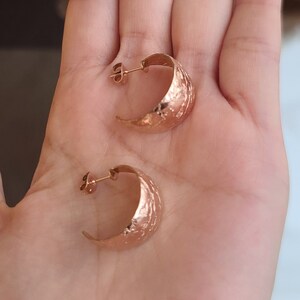 May include: A pair of rose gold hoop earrings with a textured, hammered finish. The earrings are crescent-shaped and have a post closure. The earrings are displayed on a light skin tone.