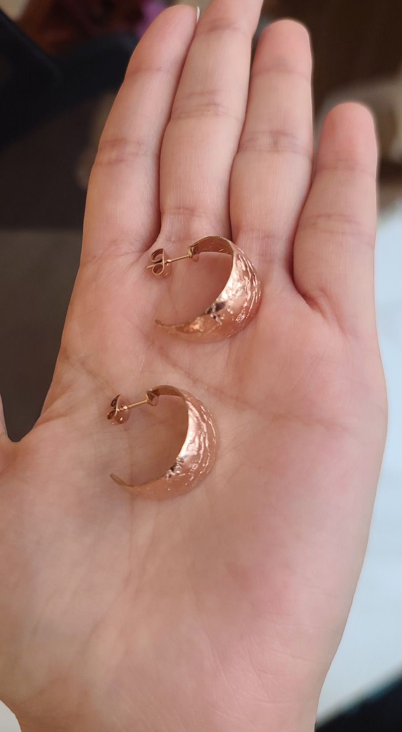 May include: A pair of rose gold hoop earrings with a textured, hammered finish. The earrings are crescent-shaped and have a post closure. The earrings are displayed on a light skin tone.