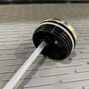 Puede incluir: Primer plano de una tapa de botella de agua negra con un borde de acero inoxidable y un anillo de silicona blanco. Una pajita de pl&aacute;stico transparente se extiende desde la tapa. La tapa est&aacute; dise&ntilde;ada para una botella de agua.