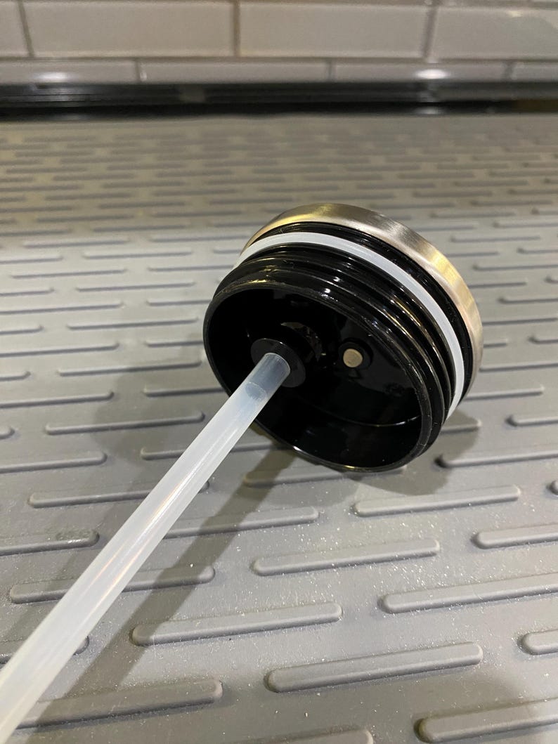 Puede incluir: Primer plano de una tapa de botella de agua negra con un borde de acero inoxidable y un anillo de silicona blanco. Una pajita de pl&aacute;stico transparente se extiende desde la tapa. La tapa est&aacute; dise&ntilde;ada para una botella de agua.