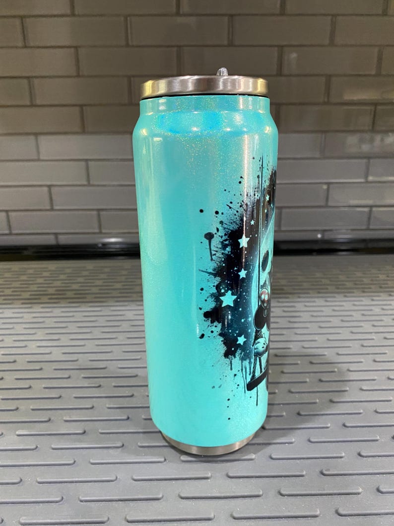 Puede incluir: Un vaso de purpurina turquesa con tapa plateada. El vaso presenta un dise&ntilde;o gr&aacute;fico negro con estrellas y salpicaduras de pintura. El vaso est&aacute; dise&ntilde;ado para parecerse a una lata de refresco.