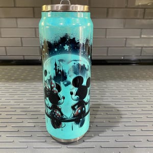 Puede incluir: Un vaso de color turquesa con tapa plateada, decorado con una silueta de Mickey y Minnie Mouse, una luna llena y un castillo. El dise&ntilde;o est&aacute; sobre un cielo estrellado, con un acabado brillante.
