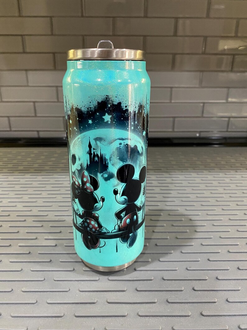 Puede incluir: Un vaso de color turquesa con tapa plateada, decorado con una silueta de Mickey y Minnie Mouse, una luna llena y un castillo. El dise&ntilde;o est&aacute; sobre un cielo estrellado, con un acabado brillante.