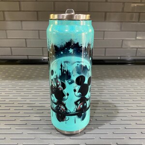 Puede incluir: Un vaso de color turquesa con tapa plateada, con un dise&ntilde;o de Mickey y Minnie Mouse frente a un castillo bajo un cielo estrellado. El vaso tiene un acabado brillante y forma de lata.