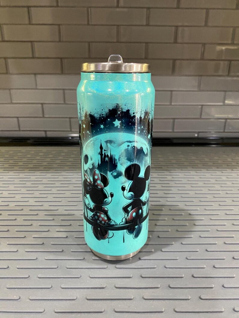 Puede incluir: Un vaso de color turquesa con tapa plateada, con un dise&ntilde;o de Mickey y Minnie Mouse frente a un castillo bajo un cielo estrellado. El vaso tiene un acabado brillante y forma de lata.