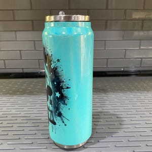 Puede incluir: Un vaso t&eacute;rmico de color turquesa con tapa plateada, dise&ntilde;ado para parecerse a una lata de refresco. El vaso presenta un gr&aacute;fico en blanco y negro con detalles de estrellas y salpicaduras de pintura. El vaso mide aproximadamente 15 cm de alto.