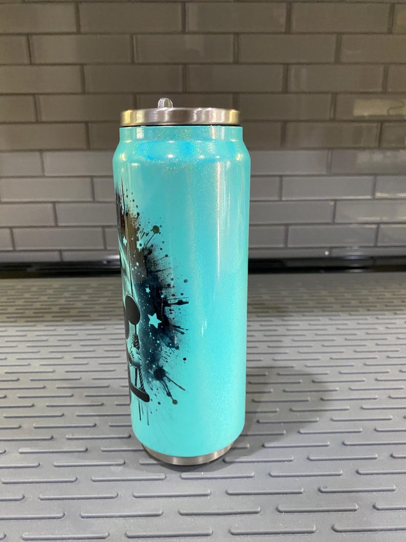 Puede incluir: Un vaso t&eacute;rmico de color turquesa con tapa plateada, dise&ntilde;ado para parecerse a una lata de refresco. El vaso presenta un gr&aacute;fico en blanco y negro con detalles de estrellas y salpicaduras de pintura. El vaso mide aproximadamente 15 cm de alto.