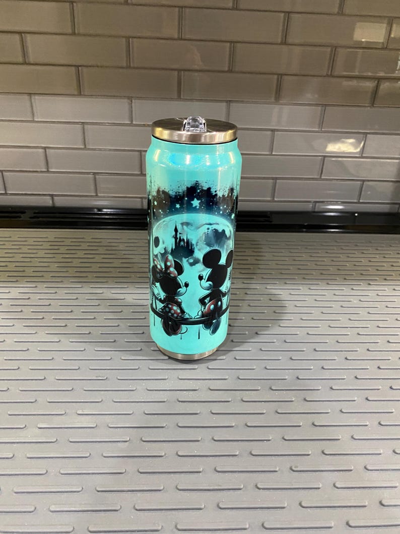 Puede incluir: Un vaso de acero inoxidable de color turquesa con tapa plateada y cierre abatible transparente. El vaso presenta una ilustraci&oacute;n en blanco y negro de Mickey y Minnie Mouse, con un castillo y un dise&ntilde;o de cielo estrellado.
