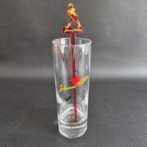 May include: A clear glass with a red and yellow stirrer featuring the Johnnie Walker logo. The stirrer has a figure of a man walking with a cane. The Johnnie Walker logo is also printed on the glass.
