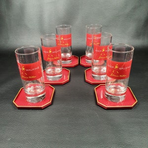 May include: Six clear glass tumblers with red bands featuring the text "KEEP WALKING" and the Johnnie Walker logo. The tumblers sit on red octagonal coasters with a gold trim. The set is arranged on a dark surface.