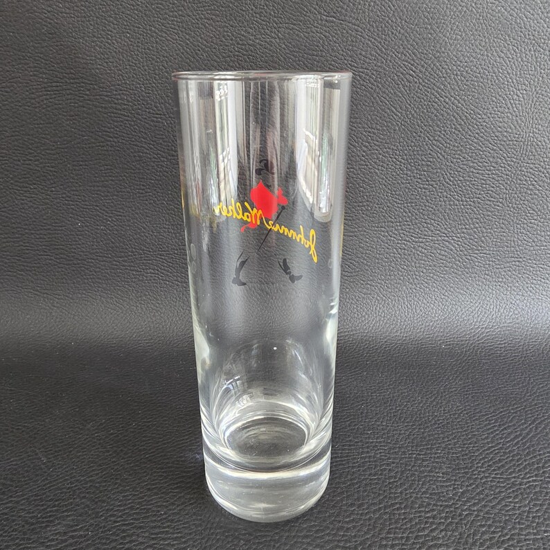 May include: A clear, tall glass with a slightly flared rim. The glass features a red and yellow logo with the text "Johnnie Walker" in a cursive font. The logo includes a silhouette of a man with a top hat.