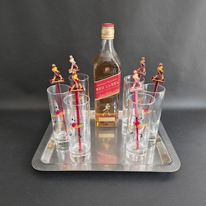 May include: A silver serving tray holds a bottle of Johnnie Walker Red Label whisky, six clear highball glasses, and six red and yellow cocktail stirrers. The glasses and stirrers feature a logo. The bottle is centered.