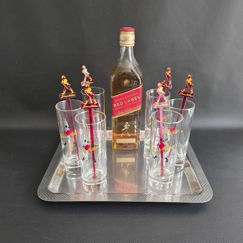 May include: A silver serving tray holds a bottle of Johnnie Walker Red Label whisky, six clear highball glasses, and six red and yellow cocktail stirrers. The glasses and stirrers feature a logo. The bottle is centered.