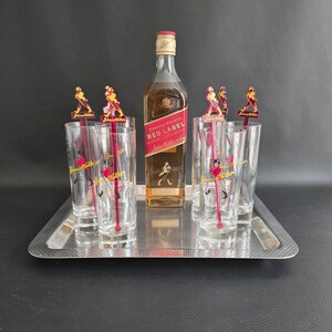 May include: A silver serving tray holds a bottle of Johnnie Walker Red Label whisky, six tall glasses, and cocktail stirrers. The glasses and stirrers feature a red and gold logo. The bottle is amber-coloured.